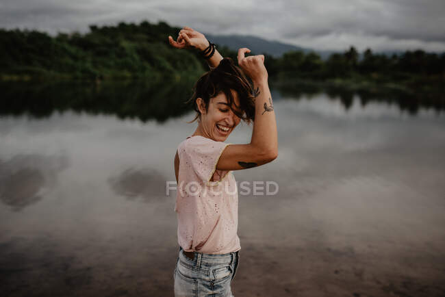 Vista lateral de excitada joven hembra sonriendo y bailando con los ojos cerrados y las manos levantadas cerca del estanque con aguas tranquilas en el campo - foto de stock
