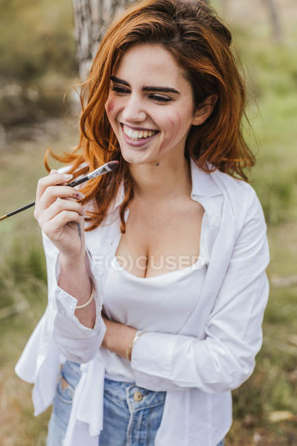 Привлекательная женщина с кисточкой улыбается и смотрит в сторону, проводя время и живопись на природе — стоковое фото