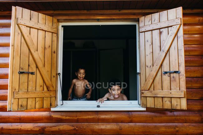 Zwei hemdlose afrikanisch-amerikanische Brüder blicken aus dem offenen Fenster eines Holzhauses, während sie zu Hause zusammen Spaß haben — Stockfoto