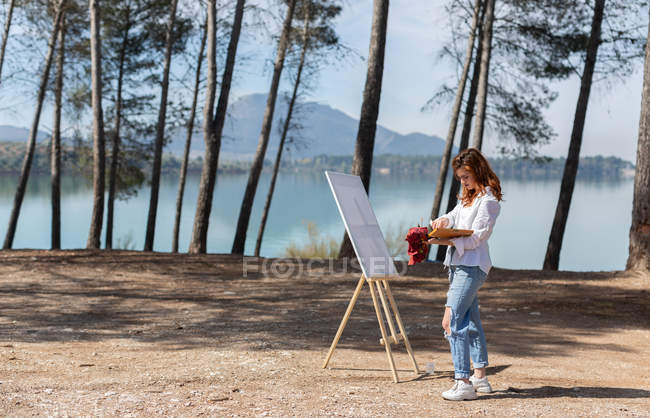 Side view of woman standing near easel and painting while spending time near calm lake on sunny day in countryside — Stock Photo