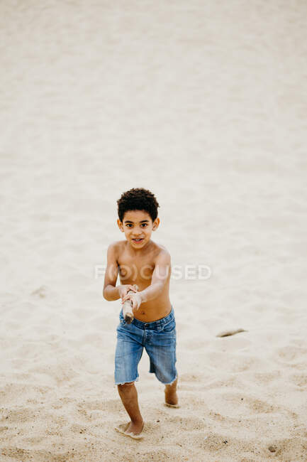 Funny African American boy with stick playing on sandy shore near sea — Stock Photo