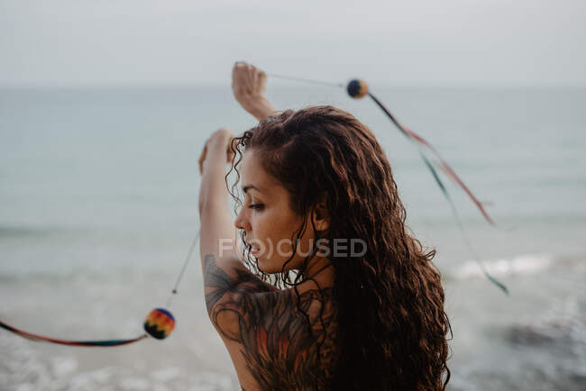 Back view of young tattooed female in swimwear swinging threads with balls while dancing near stormy sea — Stock Photo
