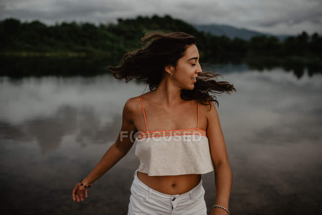 Вид збоку збуджена молода жінка посміхається і танцює із закритими очима і піднімає руки біля ставка зі спокійною водою в сільській місцевості — стокове фото