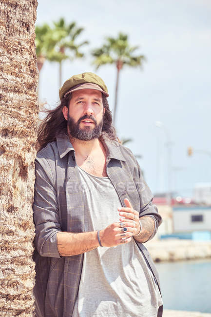 Стильний бородатий чоловік з довгим волоссям, що спирається на пальму — стокове фото
