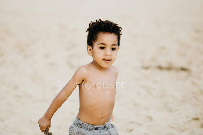 Funny African American boy with stick playing on sandy shore near sea — Stock Photo