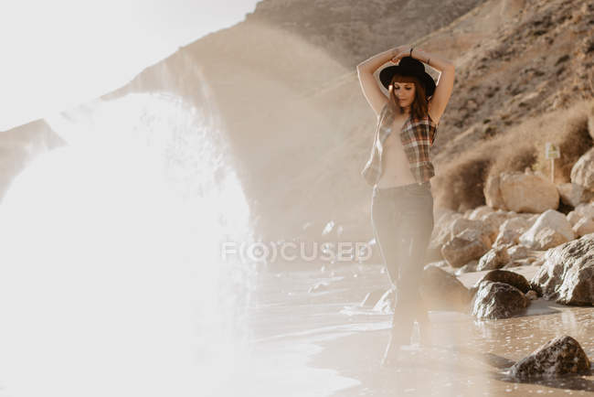 Приваблива жінка з незакріпленою картатою сорочкою, що йде біля морської води на скелястому узбережжі проти гір у сонячний день у сільській місцевості — стокове фото