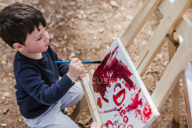 Милий маленький хлопчик малює абстрактну картину на мольберті, проводячи час у сільській місцевості — стокове фото