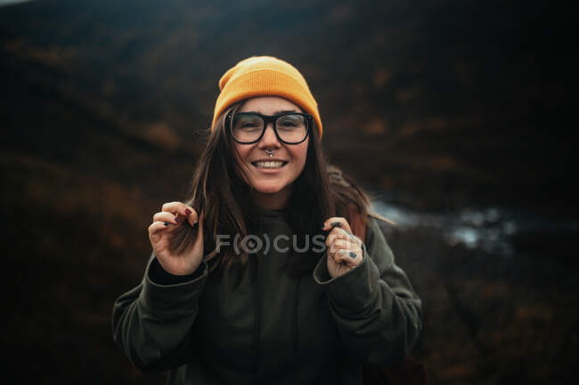 Красива молода жінка в модному капелюсі і окулярах посміхається і дивиться на камеру під час подорожі по сільській місцевості — стокове фото
