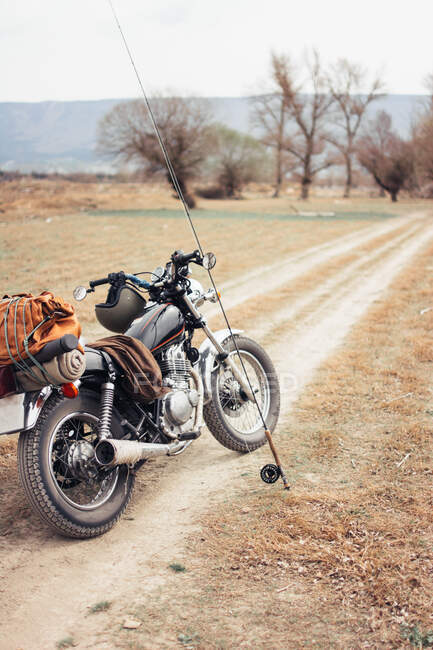Fishing pole and motorcycle located on narrow countryside road in dry field during trip in nature - foto de stock