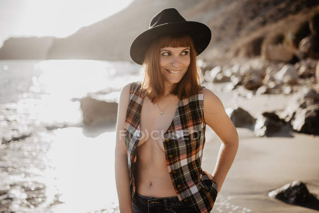Attractive female with unbuttoned checkered shirt walking near sea water on rocky coast against mountains on sunny day in countryside — Stock Photo