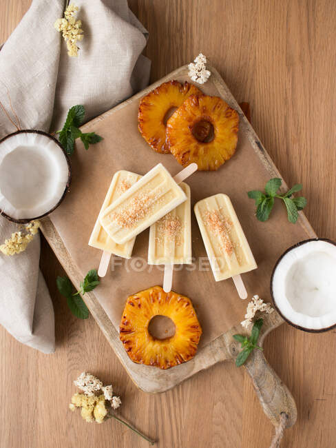 Rodajas de piña fresca y mitades de coco maduro con menta colocadas alrededor de deliciosos helados a bordo cerca de la servilleta contra la mesa de madera - foto de stock