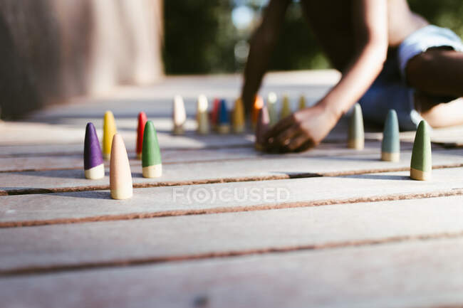 Irreconocible niño afroamericano sin camisa sentado en la superficie de madera y jugando con conos de colores en el día soleado - foto de stock