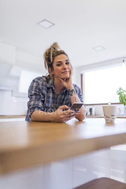 Schöne junge Frau, die zu Hause frühstückt und Spaß hat und mit dem Smartphone chattet — Stockfoto