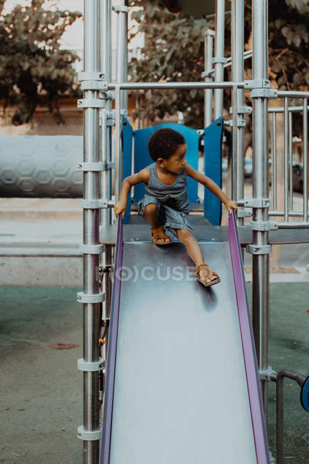 Adorabile bambino afroamericano in abito casual sorridente e seduto sullo scivolo sul parco giochi sulla strada della città — Foto stock