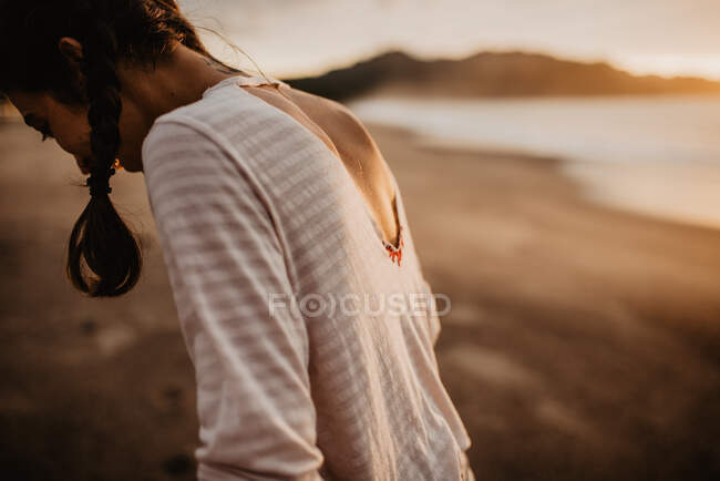 Vista trasera de la joven hembra en traje casual de pie en la playa de arena hacia el mar durante la puesta del sol en la naturaleza - foto de stock