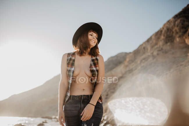 Atractiva hembra con camisa a cuadros desabotonada caminando cerca del agua de mar en la costa rocosa contra las montañas en un día soleado en el campo - foto de stock