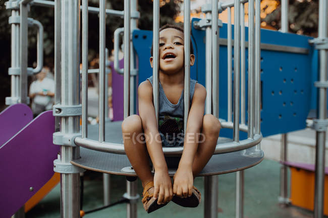Chico afroamericano con cara divertida mirando a la cámara mientras está sentado detrás de la rejilla en el patio de juegos en el parque - foto de stock