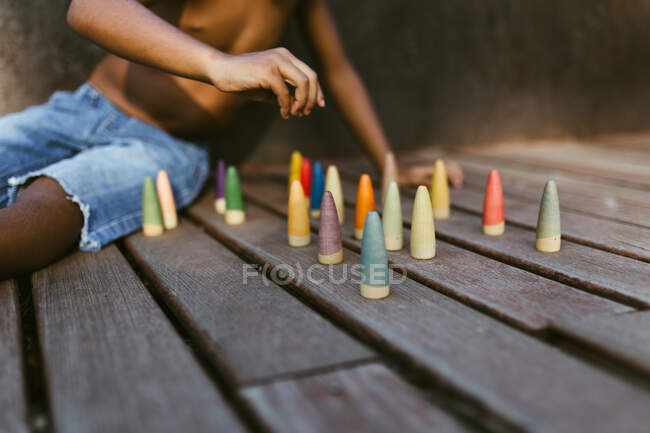 Unerkennbarer, hemdloser afroamerikanischer Junge sitzt auf einer Holzfläche und spielt an sonnigen Tagen mit bunten Zapfen — Stockfoto