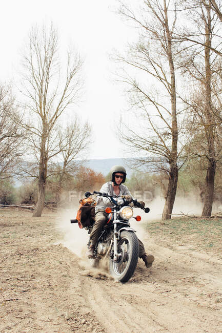 Мужчина в шлеме на быстром мотоцикле по пыльной сельской дороге возле лиственных деревьев в природе — стоковое фото