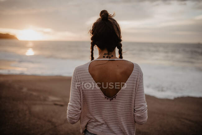 Visão traseira de jovem fêmea em roupa casual em pé na praia de areia em direção ao mar tempestuoso durante o pôr do sol na natureza — Fotografia de Stock