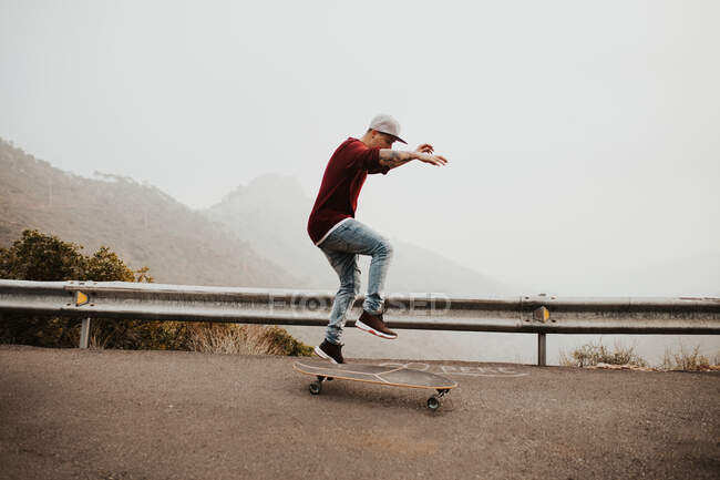 Vista lateral do jovem montando prancha longa na estrada remota da montanha e pulando contra a paisagem nebulosa — Fotografia de Stock