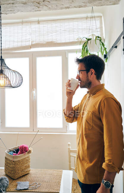 Hombre adulto disfrutando de té caliente fresco y mirando hacia otro lado mientras está sentado en la mesa cerca de agujas de punto e hilo - foto de stock