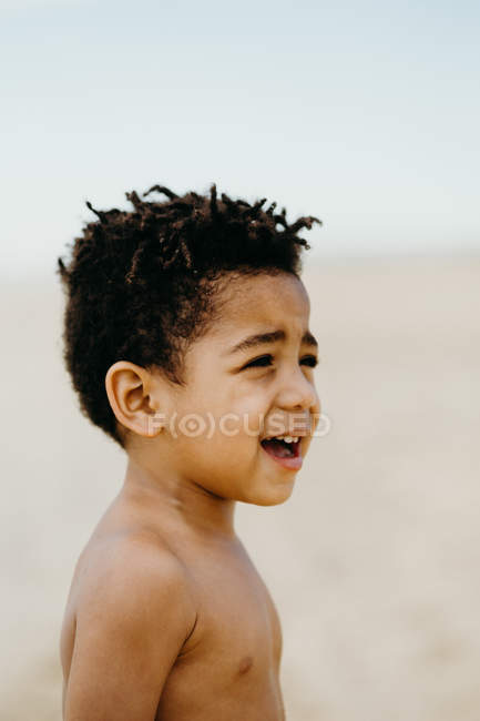 Vista lateral de un adorable niño afroamericano sin camisa mirando hacia otro lado mientras está de pie sobre un fondo borroso de playa - foto de stock