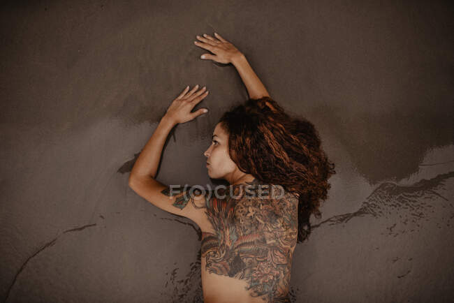 Mulher nua com costas tatuadas e ombro deitado na areia molhada na praia — Fotografia de Stock