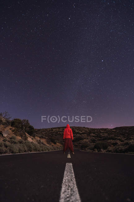 Viajero con chaqueta con capucha roja de pie en la carretera vacía por la noche - foto de stock