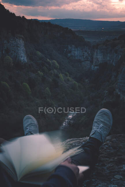 View to small river in canyon and legs of person with book sitting on the edge — Stock Photo