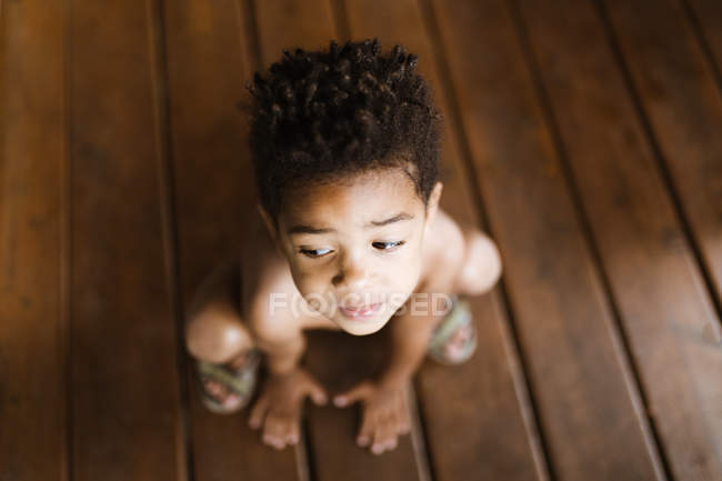 Сверху безрубашечный афроамериканец смотрит в сторону, сидя дома на деревянном полу. — стоковое фото