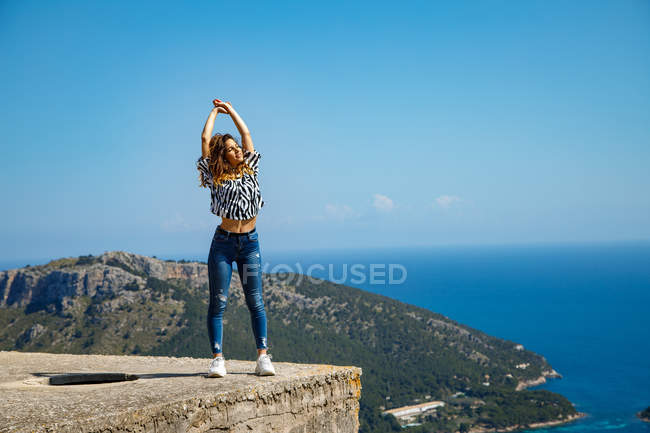 Jolie jeune femme en tenue décontractée profitant du soleil tout en se tenant sur le bloc de ciment contre ciel bleu sans nuages près de la mer — Photo de stock