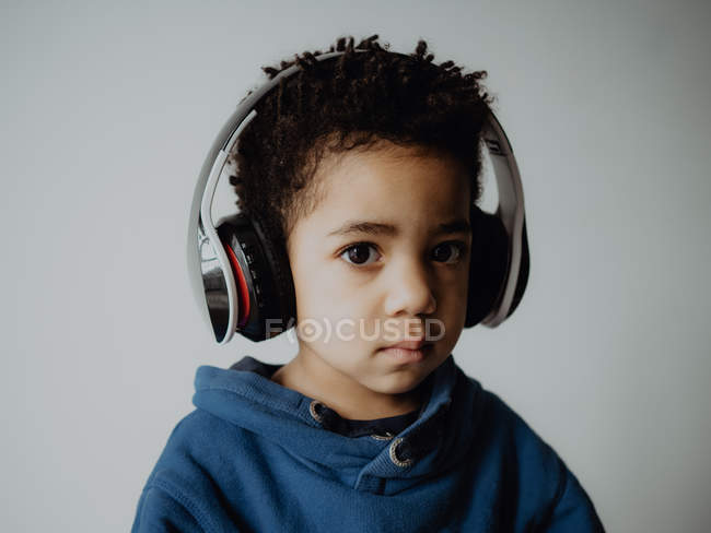 Menino afro-americano doce em camisola na moda ouvindo música em fones de ouvido, enquanto em pé contra fundo cinza — Fotografia de Stock