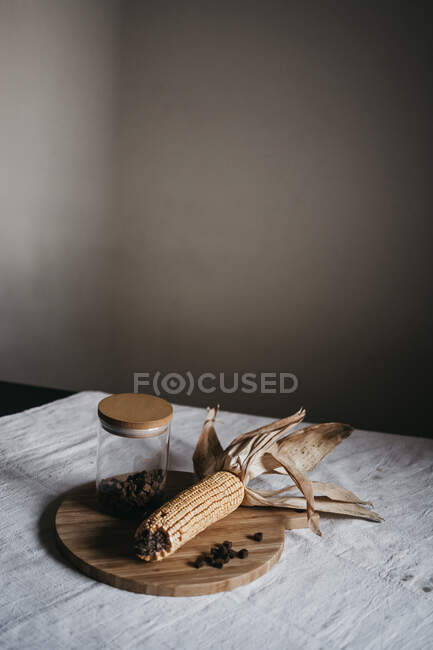 Сушеная кукуруза на початках помещена на деревянную доску возле банки с коричневыми специями на кухонном столе — стоковое фото