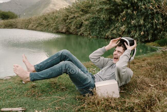 Junger Heranwachsender mit Virtual-Reality-Brille liegt mit Buch im Gras am See und lacht laut — Stockfoto