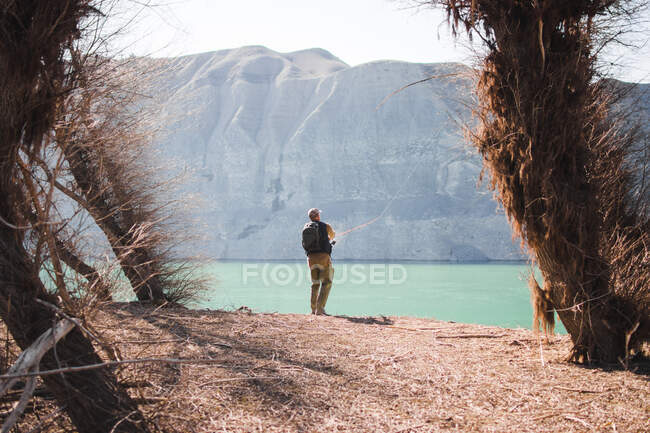 Вид сзади мужчины с рюкзаком, стоящим на берегу озера, и рыбалка в солнечный день на природе — стоковое фото