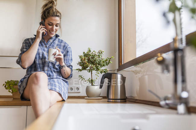 Bela e jovem mulher tomando café da manhã em casa e se divertindo, sentado na cozinha e falando ao telefone — Fotografia de Stock