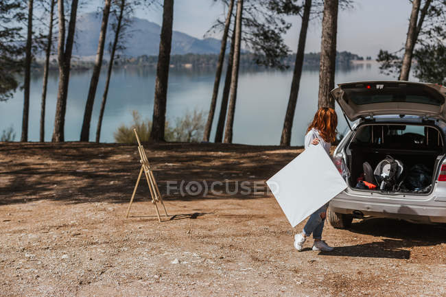 Unrecognizable woman with blank canvas standing near car with open trunk while spending time in countryside near lake — Stock Photo
