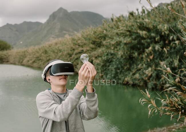 Joven adolescente jugando una simulación de realidad virtual con gafas vr de pie cerca de un lago sosteniendo una bombilla iinovación y concepto de energía - foto de stock