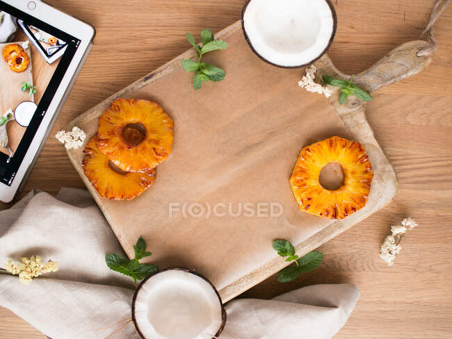 Foglie di menta fresca poste su tavola di legno vicino a fette di ananas dolce e metà di gustoso cocco su tavolino di legno vicino a tavoletta e tovagliolo — Foto stock