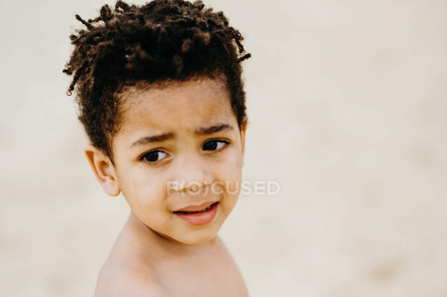 Vista lateral de un adorable niño afroamericano sin camisa mirando hacia otro lado mientras está de pie sobre un fondo borroso de playa - foto de stock