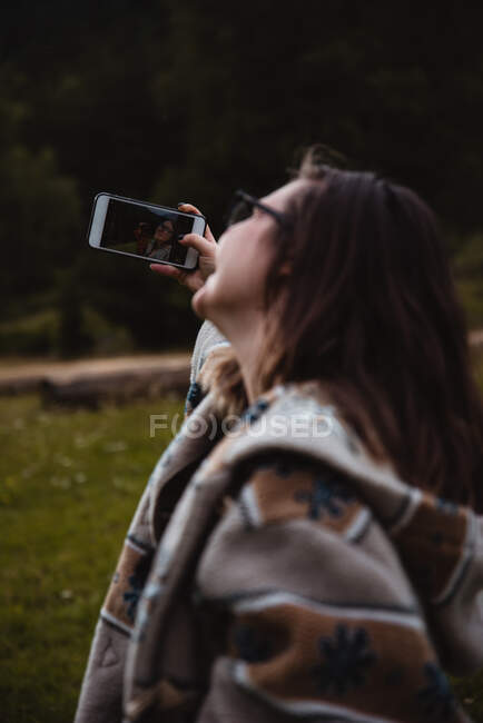 Вид сбоку на молодую женщину, использующую смартфон, чтобы сделать селфи, проводя время в величественной сельской местности — стоковое фото