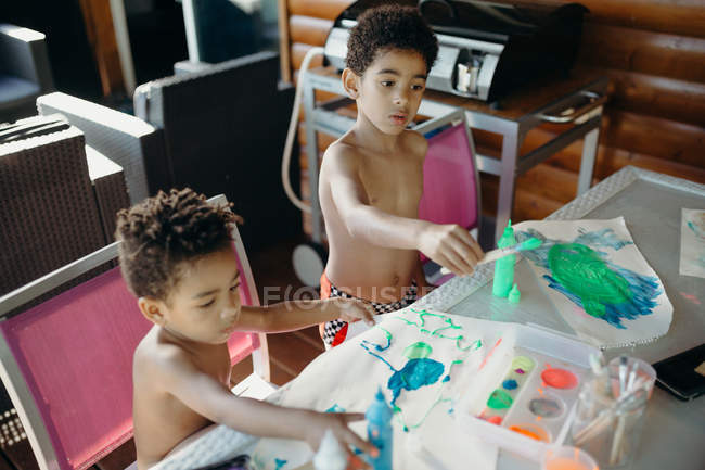 Dois meninos afro-americanos sem camisa usando tinta brilhante para fazer imagens abstratas na mesa em casa — Fotografia de Stock