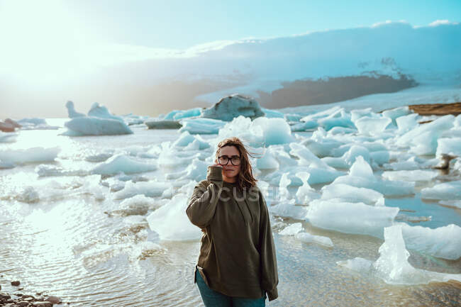 Красивая молодая женщина в повседневной одежде смотрит в камеру, стоя на берегу против бурлящей воды и тающего льда в солнечный весенний день — стоковое фото