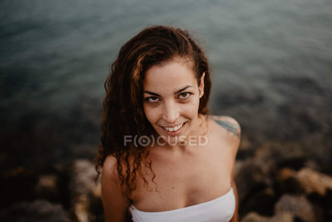 Side view of pretty young female in swimwear smiling and looking at camera while standing near calm water of sea in nature — Stock Photo