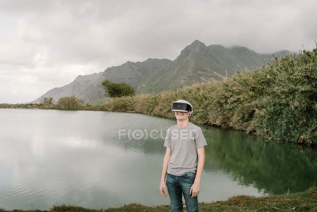 Young adolescent playing a virtual reality simulation with vr glasses standing near a lake — Stock Photo