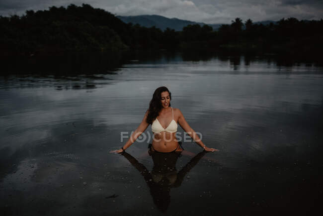 Молода жінка в купальнику стоїть у спокійній воді ставка ввечері в природі — стокове фото