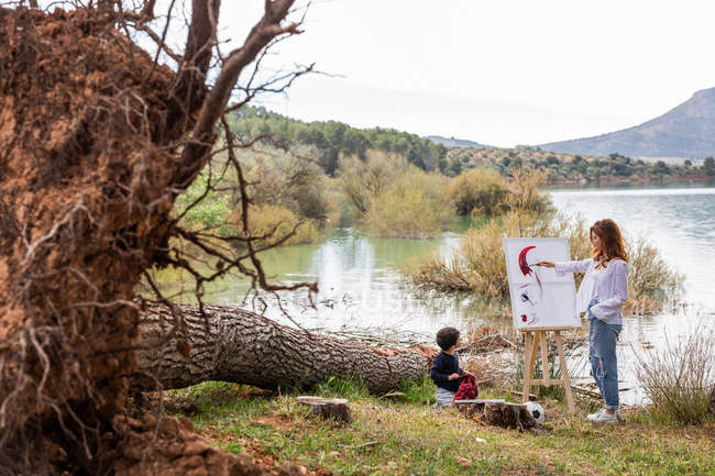 Woman with little boy painting on easel while spending time on shore of calm lake together — Stock Photo