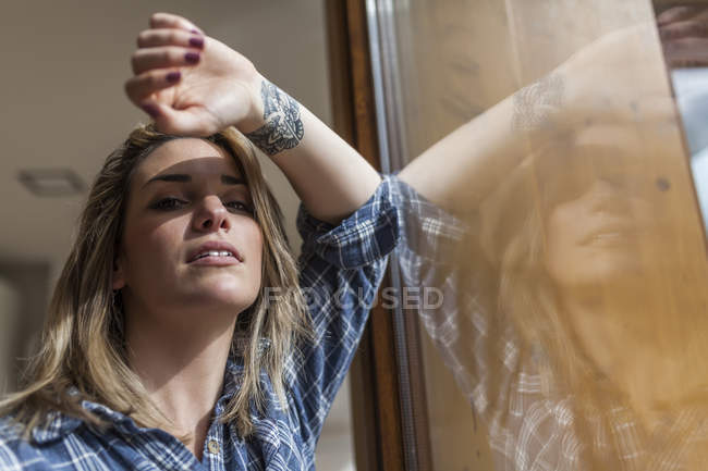 Belle et jeune femme regardant la caméra près d'une fenêtre de sa maison — Photo de stock