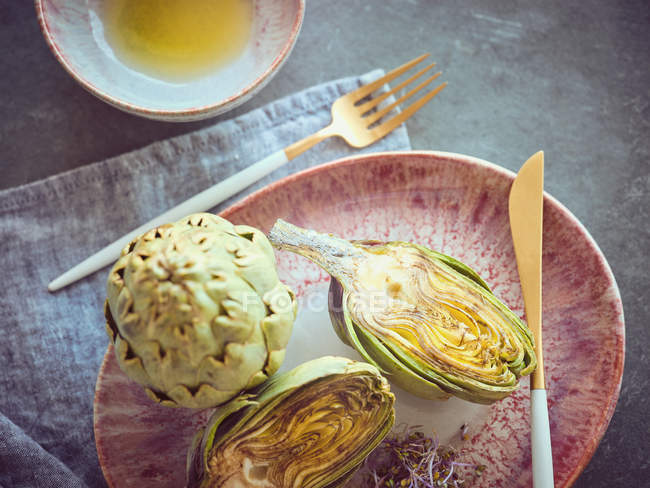 Knife and fork with whole and halved fresh artichokes on pink ceramic plate — Stock Photo